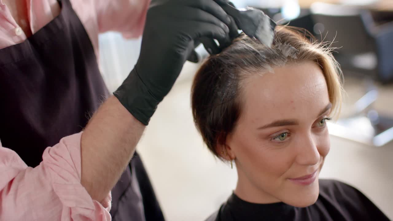 Midsection of caucasian male hairdresser dyeing happy female client's hair at salon, in slow motion