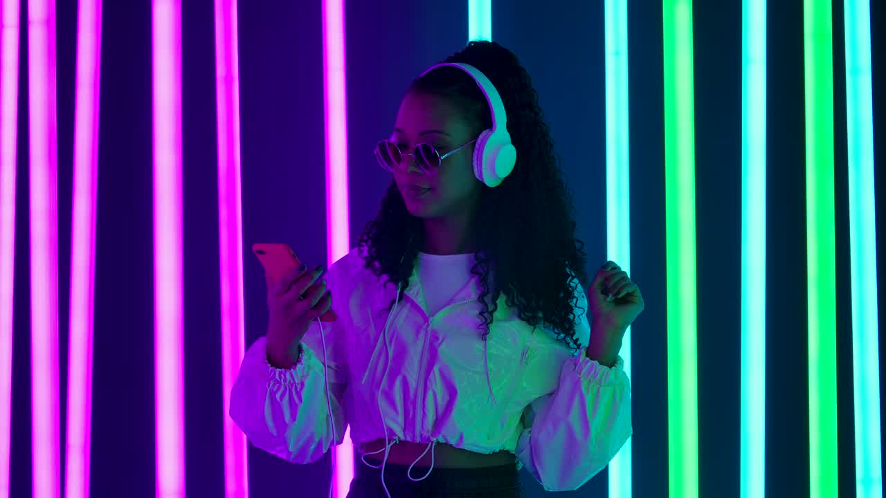 Portræt af en ung, dejlig afroamerikansk kvinde, der danser og nyder musik fra sin smartphone i store hvide hovedtelefoner. Slowmotion