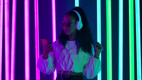 portrait of a young lovely female african american is dancing and enjoying music from smartphone in big white headphones. slow motion