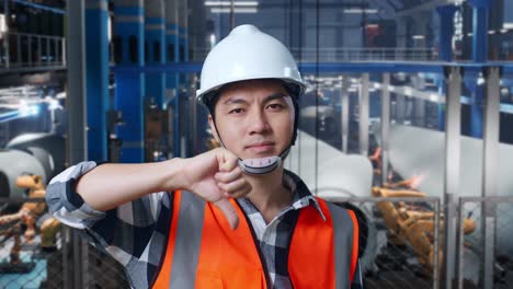 close up of asian male engineer with safety helmet standing in factory manufacture of wind turbines. showing thumbs down gesture and shaking his head while robotic arm working