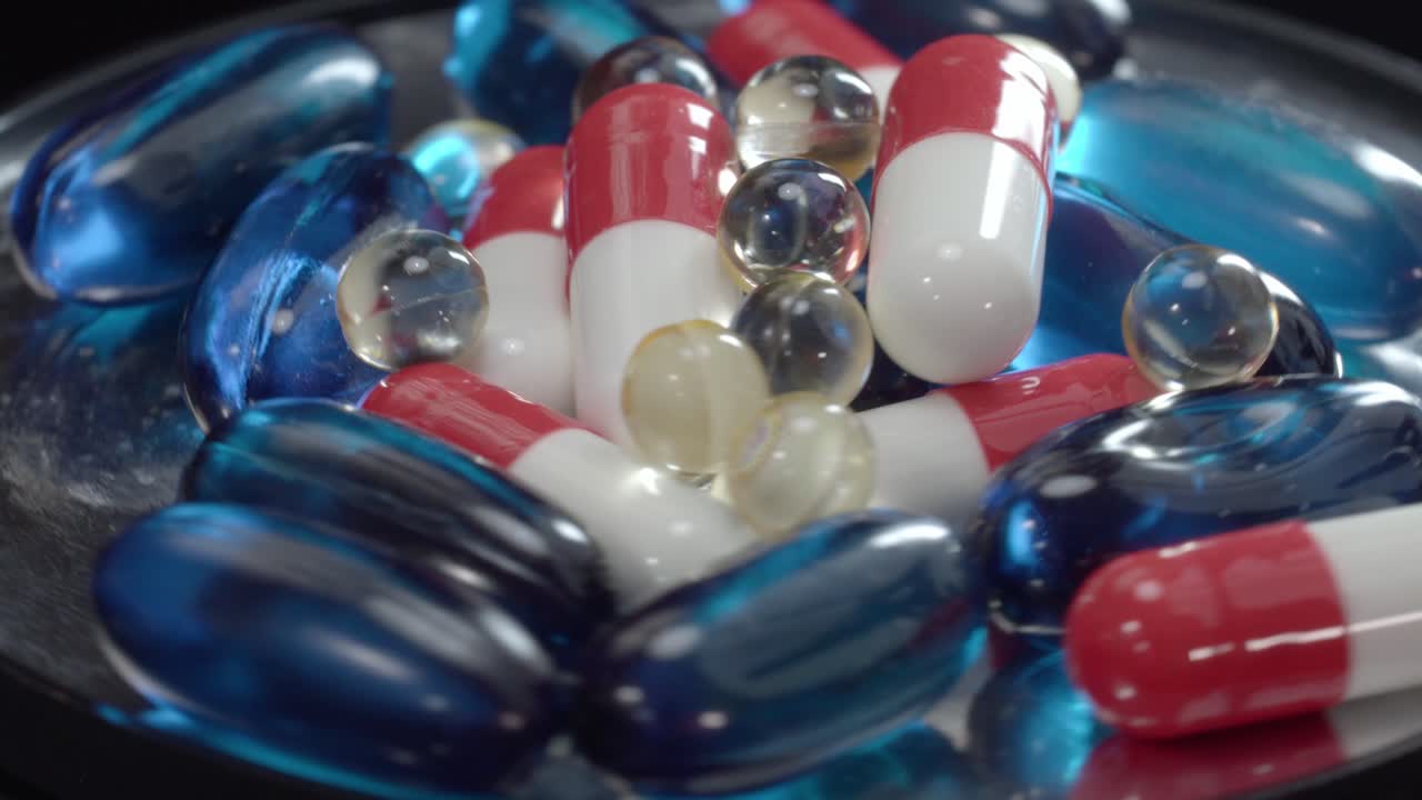 Assorted red capsules, blue gel pills and transparent softgels rotating on reflective base, studio lighting