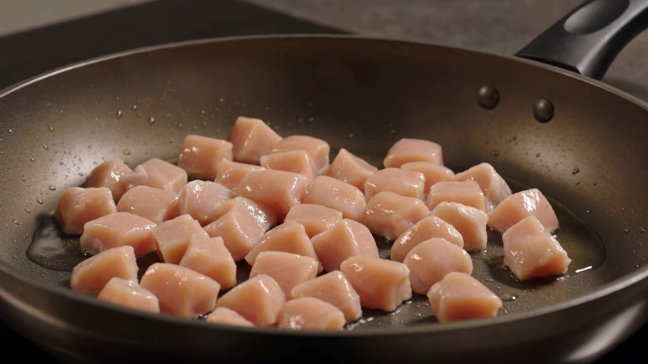家庭用コンロの上の暗いフライパンの表面に広げられた未調理の鶏肉の角切りをクローズアップ。水分を帯び、柔らかな反射があり、背景は無地のキッチン。鶏肉、生、角切り、肉。