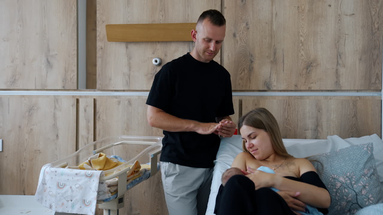 Loving husband watches his wife breastfeed their newborn. Happy parents in the hospital ward with their new baby.