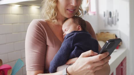 Video-of-caucasian-mother-with-newborn-baby-in-baby-carrier-using-smartphone-in-kitchen