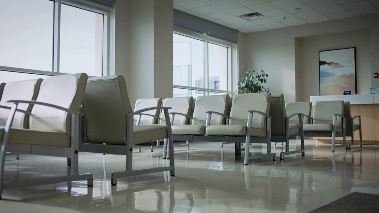 Panning camera revealing row of beige chairs in medical clinic waiting area, showing large windows