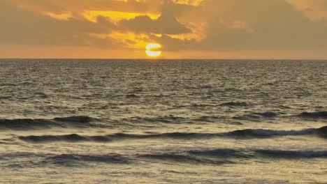 Waves-break-on-the-shore-as-sun-sets-behind-the-clouds-in-Menorca,-Spain