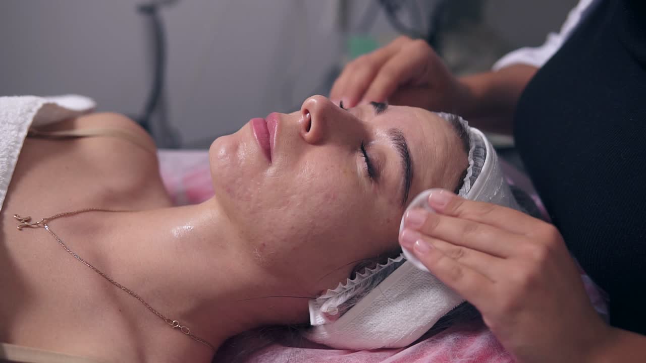 Close Up view of professional cosmetologist is cleaning woman's face using cotton sponge. Young woman is lying on the couch