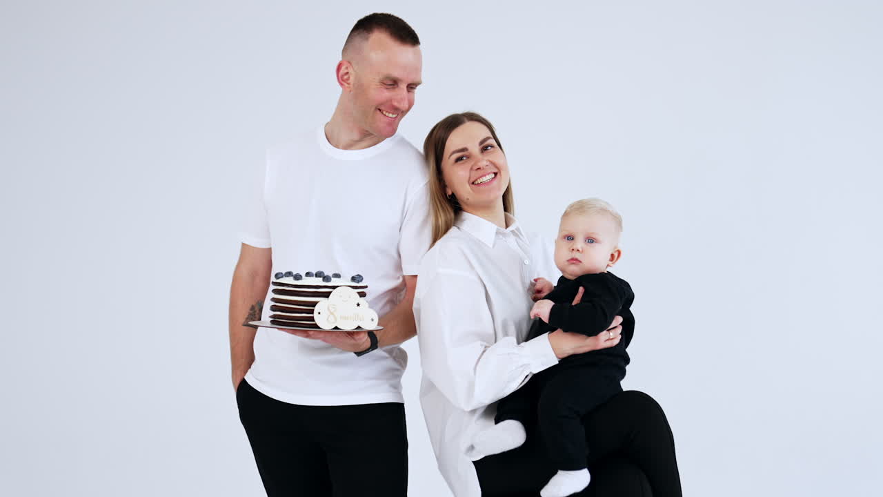Happy Caucasian family of three celebrating 8 months of their little baby. Portrait of mom, dad and son together in studio. White backdrop.