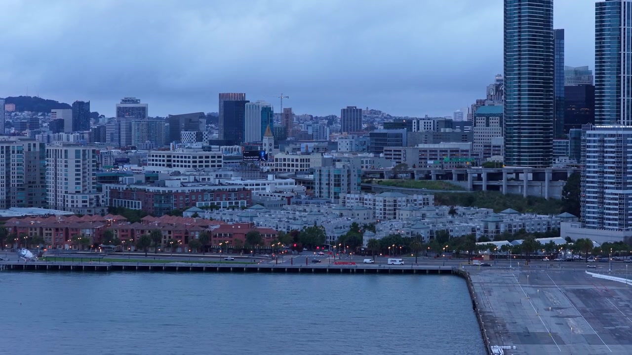 Riprese aeree catturano lo skyline e il lungomare di San Francisco al crepuscolo, con moderni grattacieli, acque calme della baia e luci urbane sotto un'illuminazione serale drammatica e nuvolosa