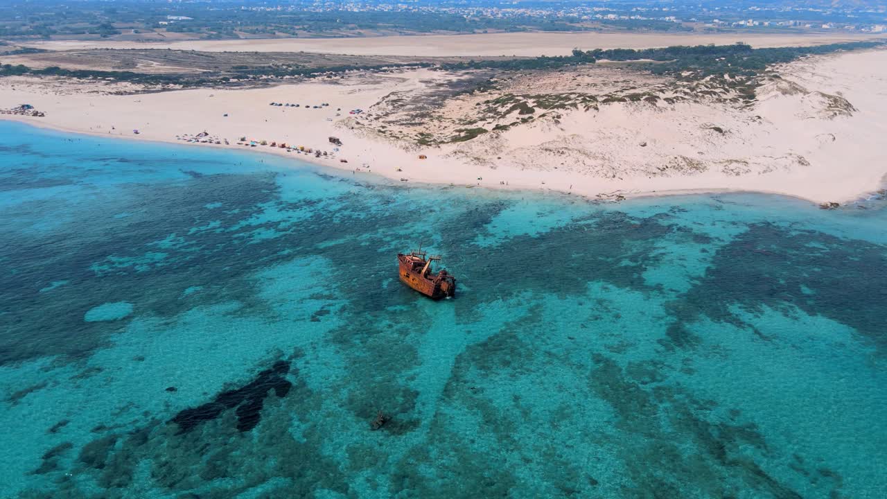 A one-of-a-kind aerial view of a weathered shipwreck lying abandoned in shallow turquoise waters, surrounded by a beach dotted with campers enjoying the coastal paradise of Tunisia's Hammam Laghzez.