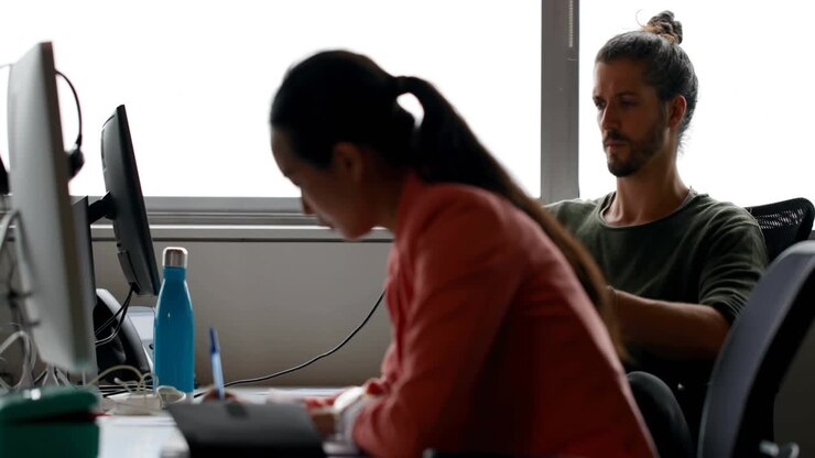 Donna che scrive e si sposta al PC per gestire le notifiche dell'ufficio mentre un collega è seduto e le icone si moltiplicano