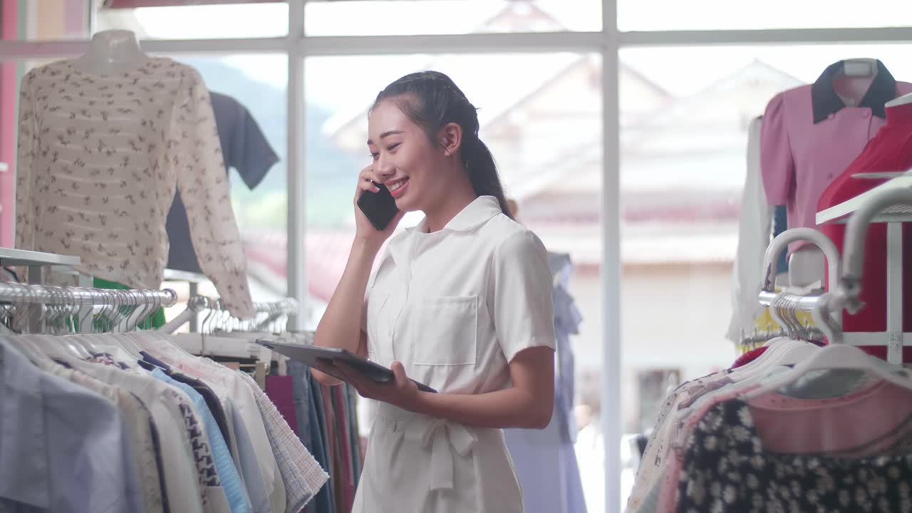 デジタルタブレットを使用してスマートフォンで話す服装店のアジア人女性オーナーがレールでストックをチェックしています