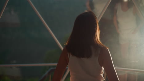 woman walks to reflection in glass under dome. tourist rests in spherical hut at modern glamping in evening. lady near window with mirror surface