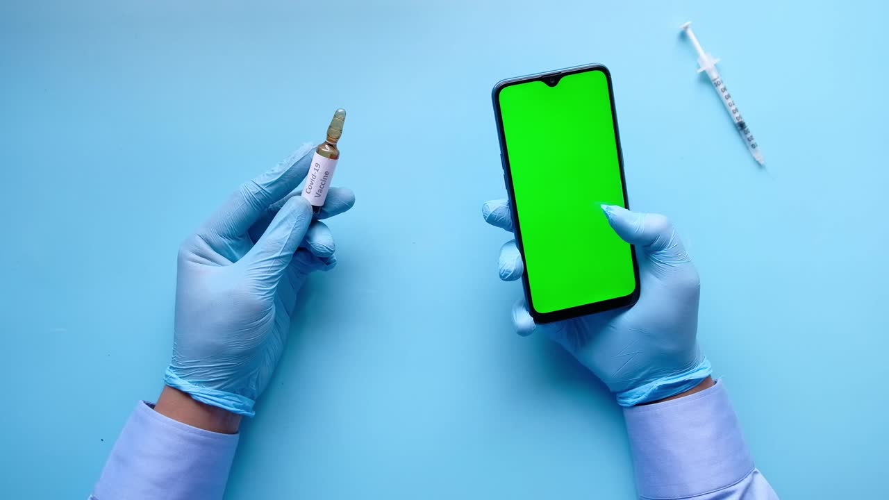 mano con guantes de látex sosteniendo una ampolla de vidrio vacuna y teléfono inteligente en azul