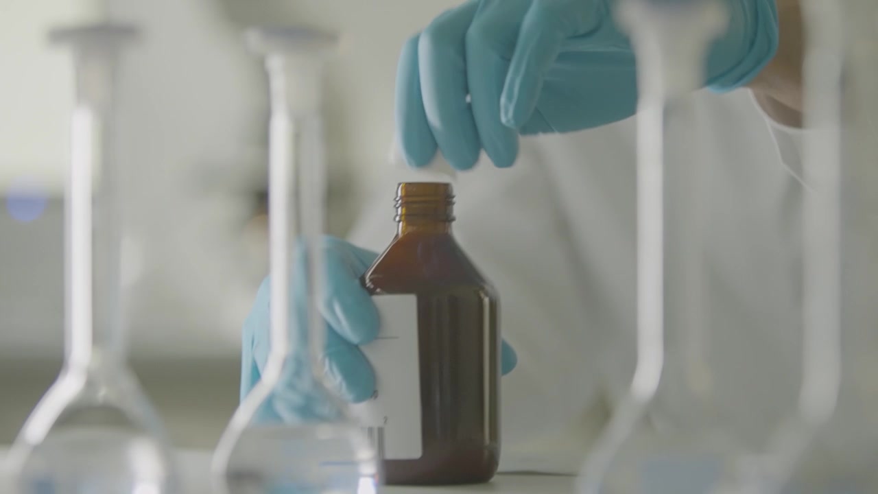 Mani guantate che aprono una bottiglia da laboratorio marrone tra vetreria in un laboratorio luminoso.