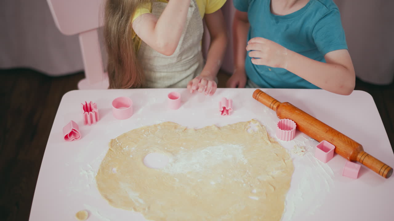 Children playing during baking session, boy using pink cutter on dough while girl removes decorative cutouts, capturing playful moment with flour scattered on table and rolling pin placed nearby