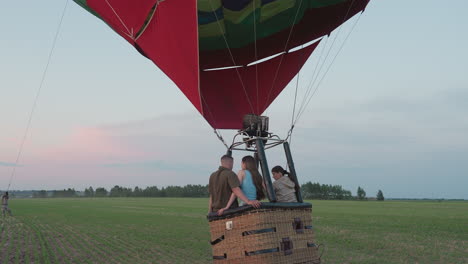 man in background pulling hot air balloon tether while woman grips rope onboard basket with passengers flying low over cultivated farmland pastel evening sky teamwork