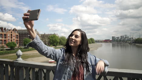 brunette latina tourist taking a selfie, posing doing victory sign with her hand, while standing on the railing of a bridge in london