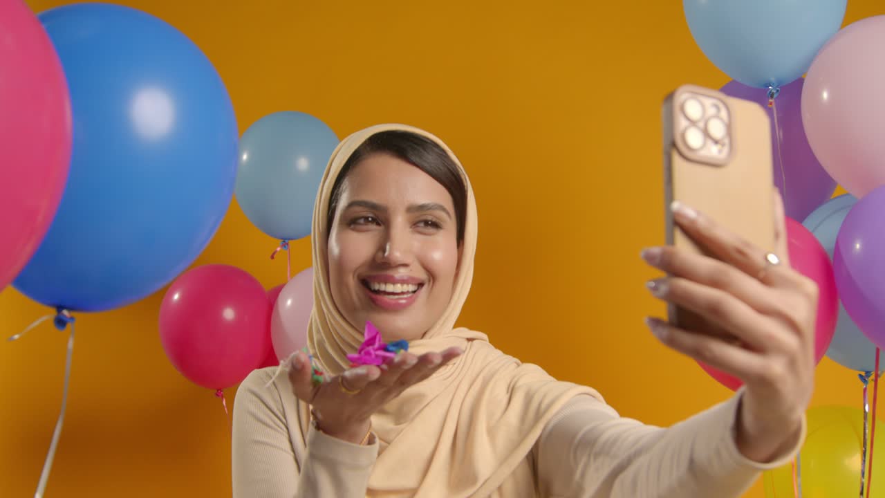 Studio Portrait Of Woman Taking Selfie Wearing Hijab Celebrating Birthday Blowing Paper Confetti Surrounded By Balloons