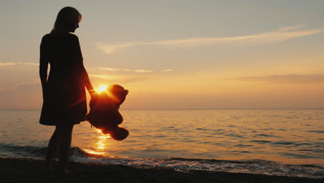 Eine-Frau-Mit-Einem-Teddybären-In-Der-Hand-Steht-Am-Meer,-Wenn-Die-Sonne-Ihre-Kindheitsträume-Versenkt