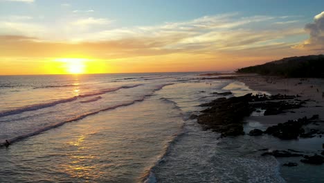 Aerial-flying-into-sunset-reflecting-in-the-water-with-waves-and-surfers-in-the-pacific-ocean-in-Tamarindo,-Costa-Rica