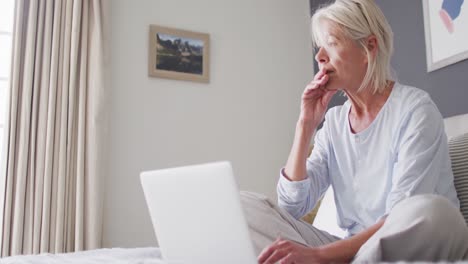 Thoughtful-senior-caucasian-woman-sitting-on-bed-in-bedroom,-using-laptop