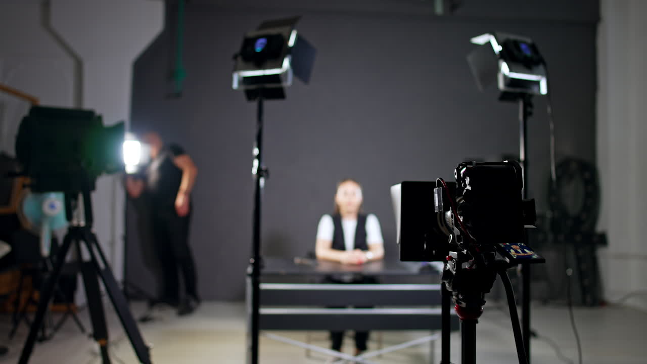 Female blogger sits at the table in studio. Cameraman walks around checking the light and equipment.