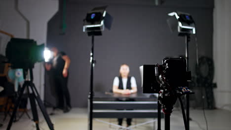 female blogger sits at the table in studio. cameraman walks around checking the light and equipment.