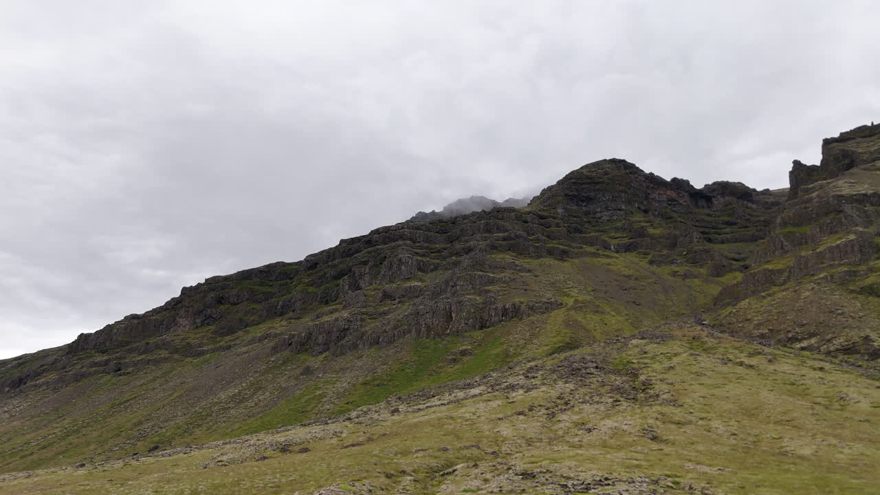 Aerial view of steep mountain cliffs in Sveitarfélagið Hornafjörður, showing layered rock formations, glacial valley textures, and sweeping rugged terrain under an overcast sky
