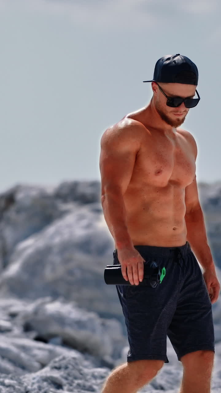 Handsome man with trained body holding flask in the mountains. Muscular athlete in shorts and sunglasses descends from the white hill in a sunny summer day. Vertical video
