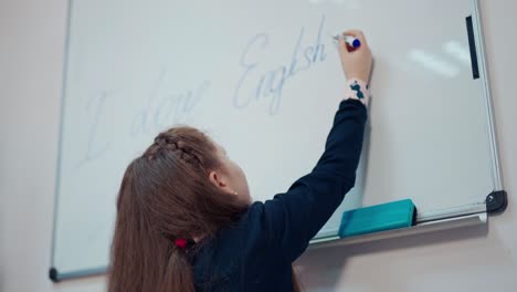 schoolgirl writes on a board. back view of a little girl in a school uniform is writing on a whiteboard with a marker. primary education concept.