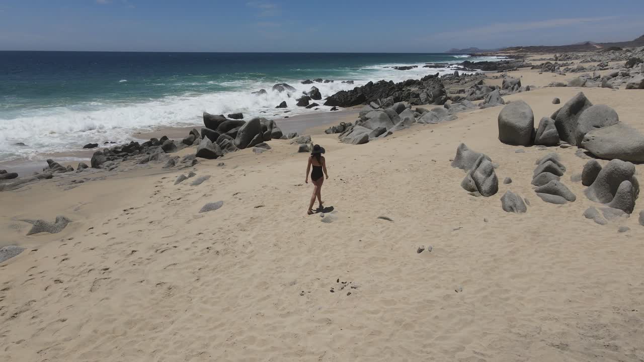 chica flaca caminando en un traje de baño negro con un sombrero negro en una playa rocosa en el sur de california