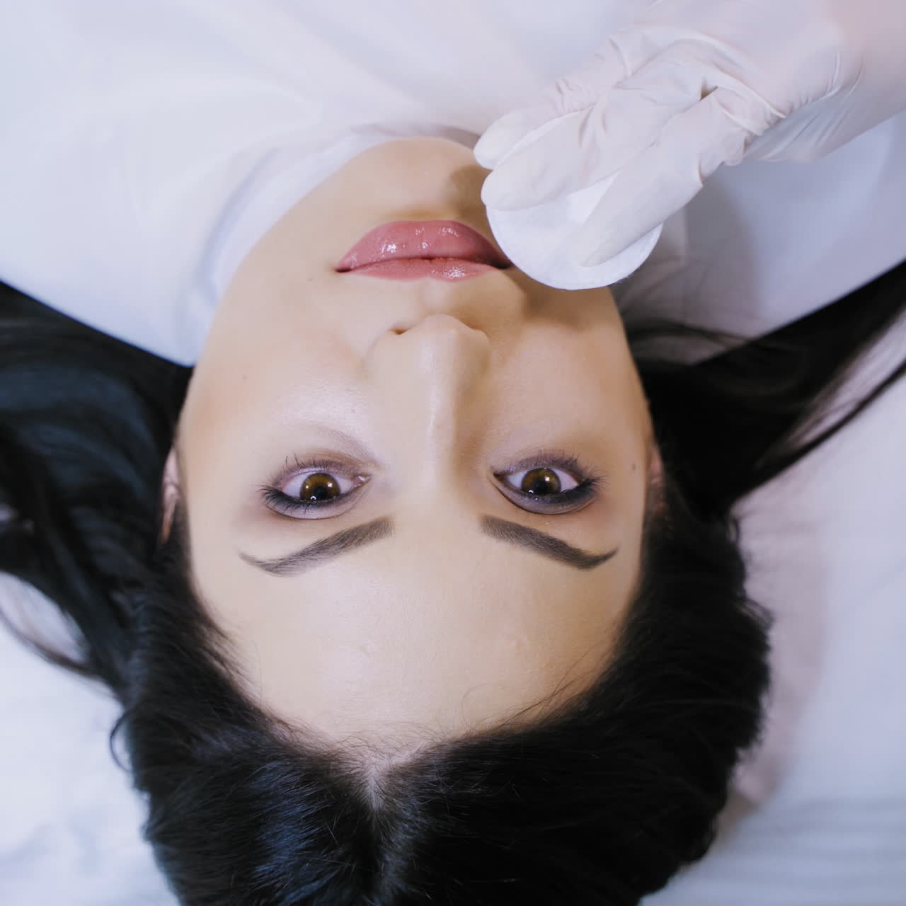 Specialist dries with a sponge lips to a woman after the permanent make-up. Top view of a beautiful female's face in a beauty salon. Slow motion
