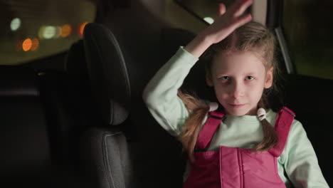 a close view of a child playfully making a hand gesture around her face while looking directly at the camera, the child appears engaged and happy, with blurred car lights visible in the background