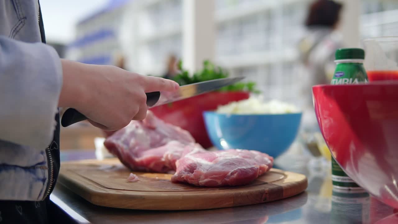 Person's hands cutting a large piece of raw meat using a butcher's knife on a wooden surface outside. Barbeque preparation. shot