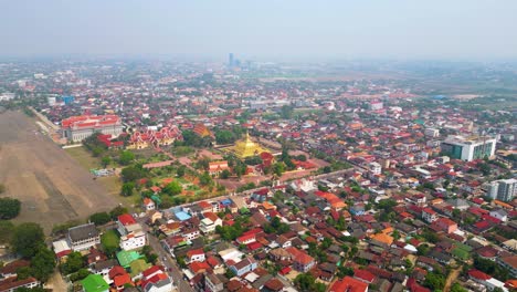 Aerial-perspective-of-Pha-That-Luang,-a-Buddhist-temple-located-in-Vientiane,-Laos,-enveloped-by-the-surrounding-local-residential-area,-portraying-the-integration-of-spirituality-and-everyday-life