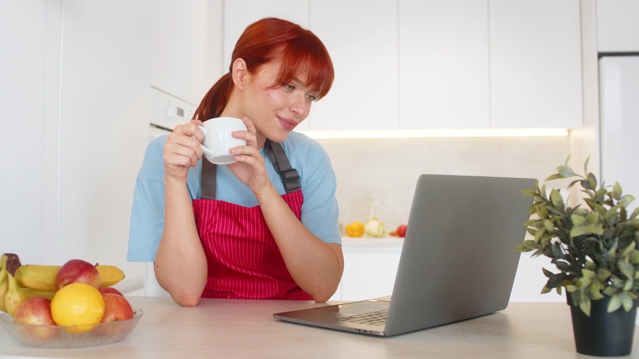 Giovane donna in cucina con il laptop risponde alle email, sorseggia caffè e gestisce le attività con concentrazione