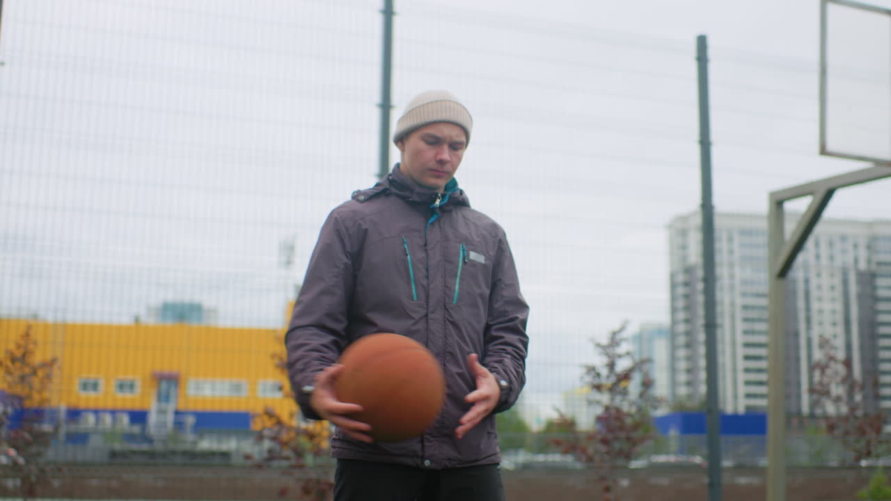 Uomo bianco con un pallone da basket in campo, che indossa un berretto di lana e una giacca impermeabile, palleggiando con ritmo costante sotto un cielo nuvoloso; canestro e recinzione a maglie a catena inquadrano la scena, sullo sfondo palazzi multipiano