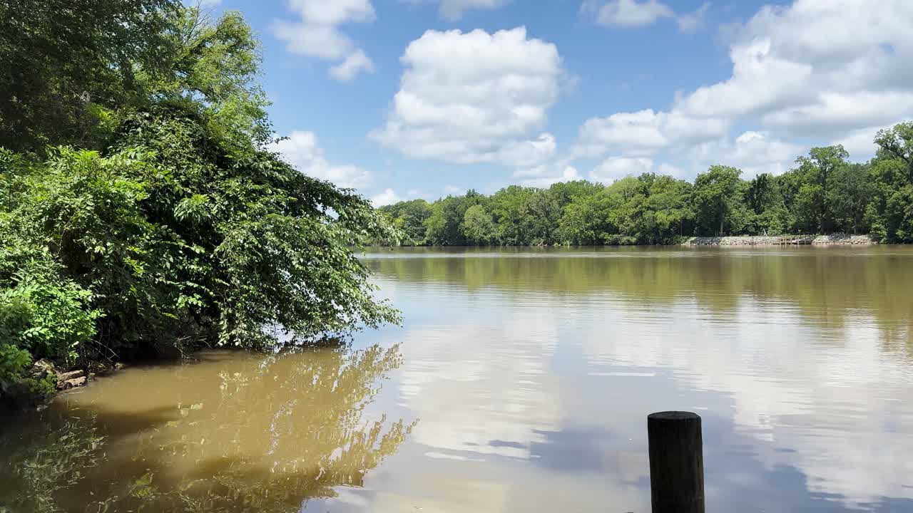 Static shot with dock post in foreground, calm water, sky and trees reflected in the Virginia River, seen from a shaded riverside