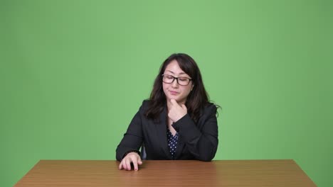 성숙하고 지루한 아시아 사업가 여성이 책상 뒤에 앉아서 생각하고 있습니다.