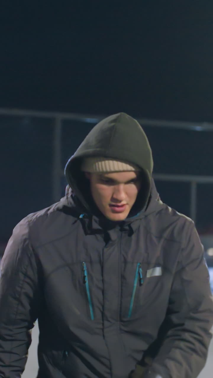 hombre con capucha con expresión enfocada durante la actividad nocturna en el campo urbano al aire libre, el fondo presenta una valla iluminada, luces amarillas y rojas vibrantes
