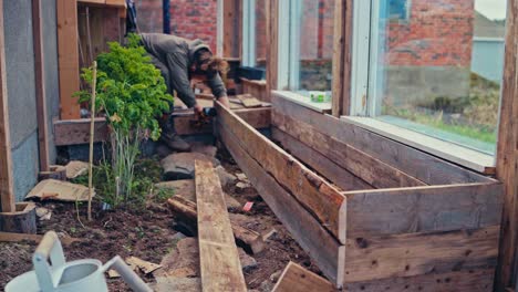 man constructing a wooden raised bed inside a small greenhouse. static shot