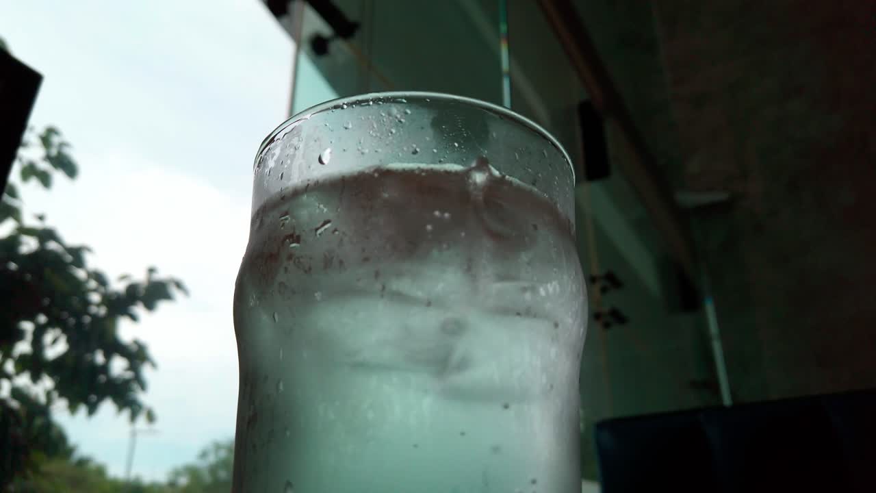Close up of a cold glass of water with ice cubes inside a café, showing condensation and urban reflections behind.