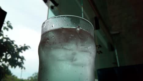 Close up of a cold glass of water with ice cubes inside a café, showing condensation and urban reflections behind.