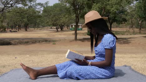 Lady-in-a-sundress-with-a-hat-sitting-and-reading-a-book-at-the-park,-static-medium-shot