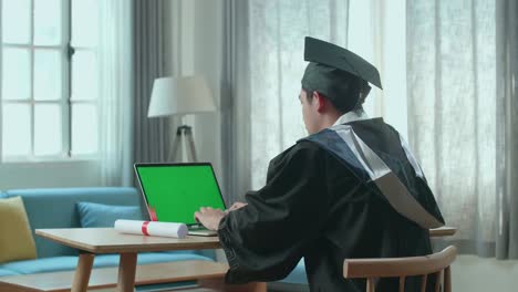 asian man wearing a graduation gown and cap use laptop computer with green mock-up screen in living room