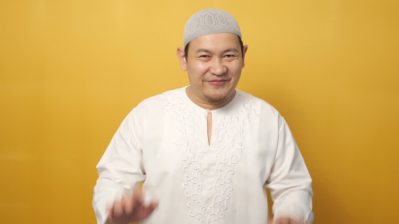 Muslimimies, jolla on hijab, katsoo kameraan hymyillen ja heiluttaen käsiään tervehdys- tai hyvästelyeleenä keltaista taustaa vasten