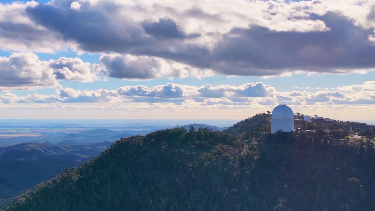 Aerial drone footage pans across a forested mountain ridge, revealing a white observatory dome under dramatic clouds and soft sunset lighting