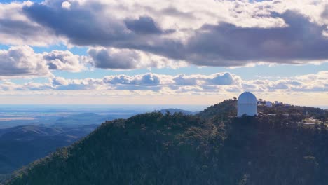 aerial drone footage pans across a forested mountain ridge, revealing a white observatory dome under dramatic clouds and soft sunset lighting