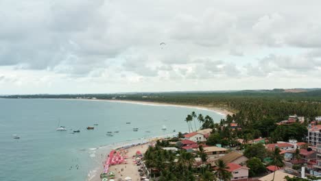 Absteigende-Luftdrohne-Weitwinkelaufnahme-Des-Hafens-Von-Porto-De-Galinhas-Oder-Hühnerhafen-Mit-Verankerten-Segelbooten,-Einem-Hängegleiter-Und-Touristen,-Die-Im-Kristallklaren-Meerwasser-In-Pernambuco,-Brasilien,-Schwimmen